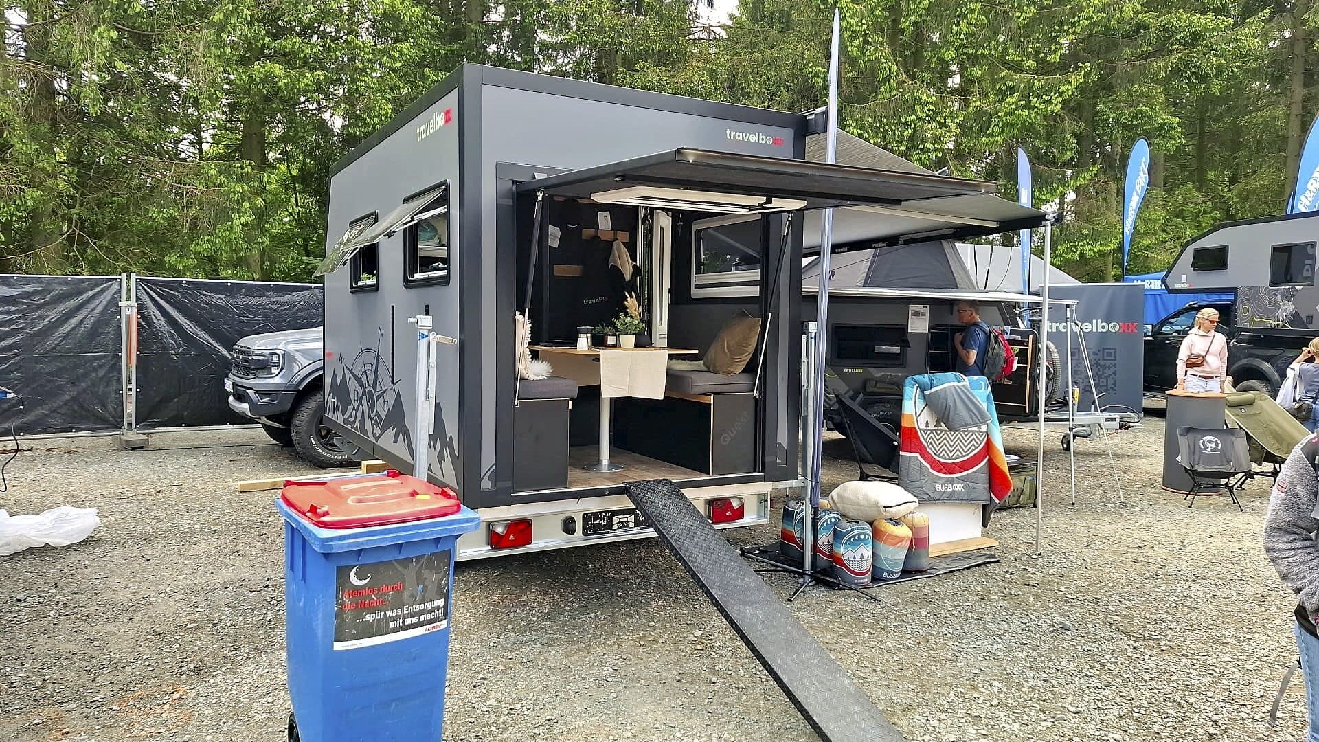 Ein moderner Pickup-Camper präsentiert sich mit geöffneter Hecktür und bietet im Inneren eine gemütliche Einrichtung mit Tisch, Sitzgelegenheiten und stilvoller Dekoration. In der Nähe befinden sich die Travelboxx Quest Campingausrüstung sowie ein blauer Mülleimer. Im Hintergrund sind Bäume und weitere Offroad-Camper oder Wohnanhänger zu sehen - ideal für alle, die auf der Suche nach einem individuellen Custom Camper für ihre nächste Abenteuerreise sind. Travelboxx - Pickup Wohnkabinen