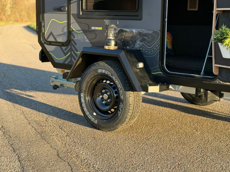 Nahaufnahme eines schwarzen Offroad-Campinganhängers: Der robuste Reifen steht auf einer sonnendurchfluteten Asphaltstraße, eine Metall-Laterne ziert den Kotflügel. Seitenteil und Einstieg des Trailers sind sichtbar – die Pickupboxx präsentiert sich als vielseitiges Allround-Talent für grenzenlose Freiheit im Outdoor-Bereich. Ob als Pickup Camper, Offroad Camper oder individuell zugeschnittener Custom Camper: Die Pickupboxx bietet maximale Flexibilität und Komfort für jedes Abenteuer abseits befestigter Wege. Travelboxx - Pickup Wohnkabinen