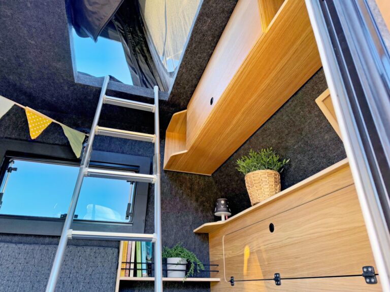 Innenraum einer modernen Pickup Camperbox: Entdecken Sie den stilvollen Ausbau eines Offroad-Campers mit maßgefertigten Holzregalen, dekorativen Zimmerpflanzen und einer stimmungsvollen Lampe. Über einen stabilen Metallleiter gelangst du bequem in den erhöhten Schlafbereich – genieße das Gefühl von grenzenloser Freiheit, während Sonnenlicht durch die großzügigen Fenster fällt. Perfekt für alle, die einen individuellen Custom Camper oder eine praktische Lösung als Pickup Wohnkabine suchen. Travelboxx - Pickup Wohnkabinen