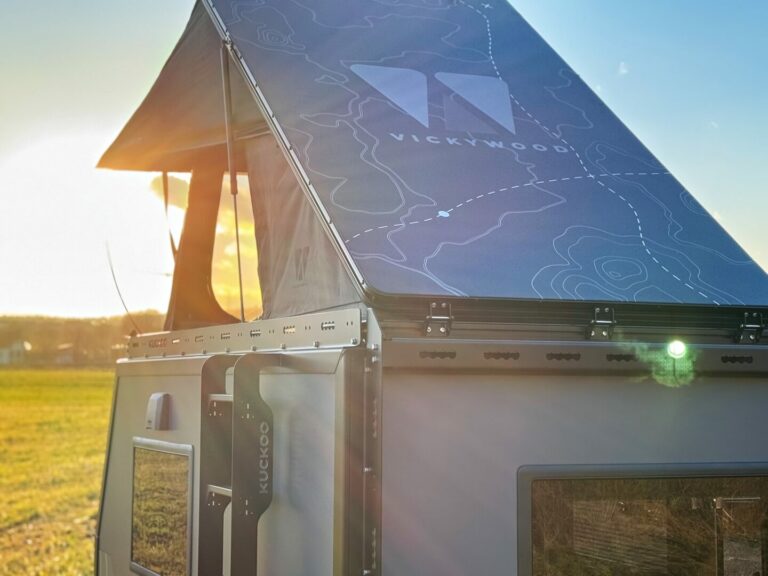 Nahaufnahme eines „VICKYWOOD“ Dachzelts, teilweise geöffnet im Sonnenlicht, montiert auf einem kompakten Offroad-Camper-Anhänger in einer grünen Wiese bei Sonnenuntergang – ideal für alle, die mit einem Pickup Camper oder Custom Camper grenzenlose Freiheit und Abenteuer in der Natur suchen. Travelboxx - Pickup Wohnkabinen