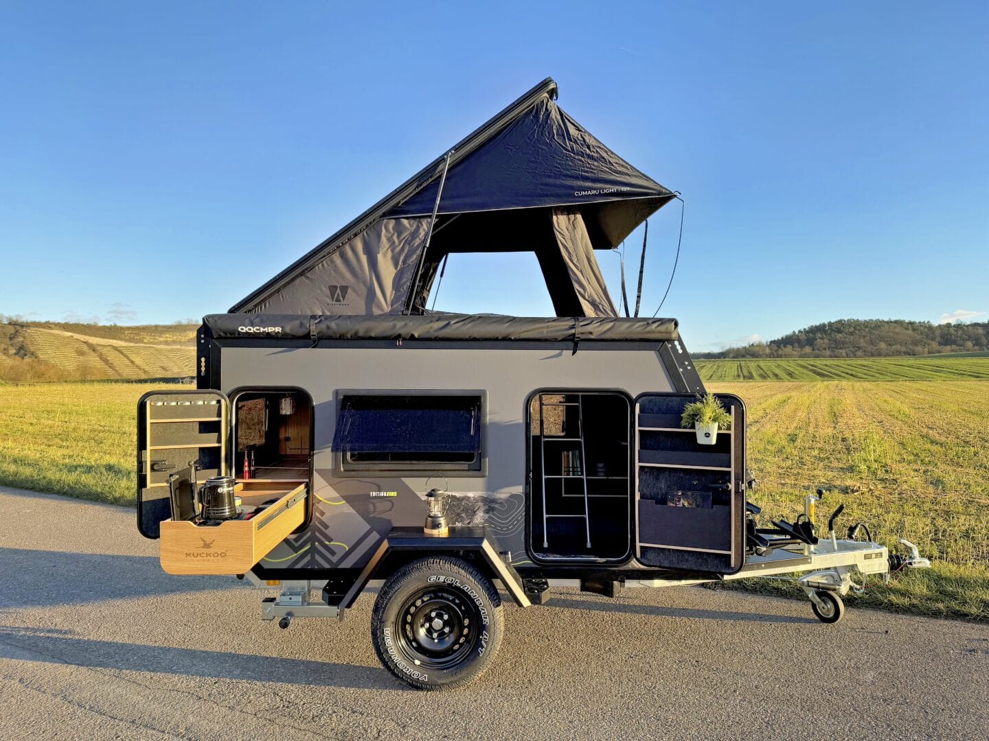 Ein kompakter, schwarzer Allround Offroad-Camper-Anhänger mit erhöhtem Aufstelldach steht auf einer ländlichen Straße neben grünen Wiesen. Die seitliche Klappe ist geöffnet und gibt den Blick auf eine durchdachte Outdoor-Küche mit Herd, Spüle und Stauraum frei. Dieser Custom Camper Trailer eignet sich ideal für abenteuerlustige Pickup-Fahrer oder alle, die nach einem flexiblen Pickup-Camper für Geländeeinsätze suchen. Travelboxx - Pickup Wohnkabinen
