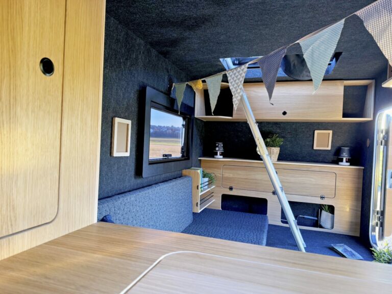 Innenraum eines modernen Pickup Campers mit hellen Holzschränken, einer grauen gepolsterten Sitzbank und kleinen Zimmerpflanzen – ideal für alle, die einen Offroad Camper oder Custom Camper suchen. Dreieckige Wimpelgirlanden hängen von der Decke und Sonnenlicht flutet den Raum – perfekt für grenzenlose Freiheit und echtes Allround-Talent unterwegs. Travelboxx - Pickup Wohnkabinen