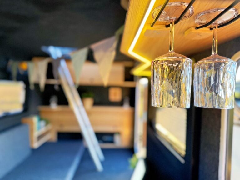 Zwei Kristallweingläser hängen kopfüber unter einem Holzregal im Inneren eines gemütlichen, modernen Pickup Campers – ein echtes Offroad-Allroundtalent. Im unscharfen Hintergrund sind eine Leiter, Wimpelgirlanden, eine Topfpflanze und maßgefertigte Holzmöbel zu erkennen. Dieses individuell ausgebaute Custom Camper Interieur kombiniert praktisches Design mit stilvollem Ambiente für Abenteuer auf und abseits der Straße. Travelboxx - Pickup Wohnkabinen