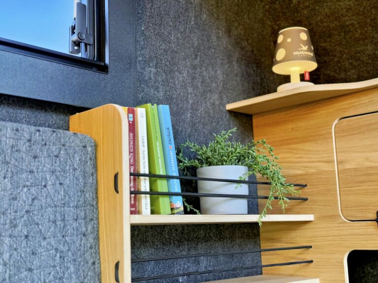 Ein kleines Regal, ein wahres Allround-Talent für deinen Pickup Camper oder Offroad Camper, bietet Platz für bunte Bücher, eine Zimmerpflanze und eine Holzlampe mit ausgestanzten Sternen. Die grauen Regalbretter und Wandflächen harmonieren perfekt mit den hellen Holzmöbeln – so entsteht in deinem Custom Camper ein gemütliches und modernes Interieur. Travelboxx - Pickup Wohnkabinen