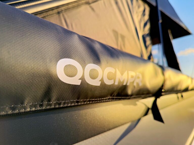 Nahaufnahme einer schwarzen Stoffabdeckung mit weißem „QQCMPR“-Schriftzug, von warmem Sonnenlicht beleuchtet – ein Symbol für grenzenlose Freiheit beim Offroad-Abenteuer. Dieses Detailbild weckt die Lust auf individuelle Pickup Camper und maßgeschneiderte Offroad Camper, perfekt für alle, die auf der Suche nach einem einzigartigen Custom Camper für ihre nächsten Outdoor-Erlebnisse sind. Im unscharfen Hintergrund zeigt sich die Natur – ideal für Abenteurer, die mit ihrem Pickup Truck Camper abseits befestigter Wege unterwegs sein wollen. Travelboxx - Pickup Wohnkabinen