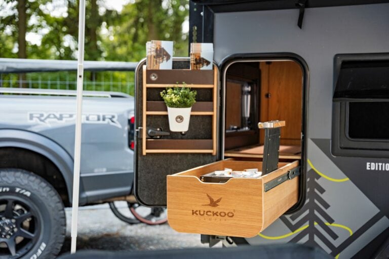 Die kompakte Travelboxx Max Camper-Küche ist ideal für alle, die einen Pickup Camper, Offroad Camper oder Custom Camper suchen. Sie verfügt über eine ausziehbare Holzschublade mit dem Schriftzug „Kuckoo“, ein kleines Pflänzchen sowie Ablagefächer im geöffneten Türbereich – optimal für Wohnanhänger- oder Tiny-House-Fans. Der Anhänger steht direkt neben einem grauen Truck und bietet somit die perfekte Lösung für flexible Camping-Abenteuer mit Ihrem Pickup oder individuellen Offroad-Camper. Travelboxx - Pickup Wohnkabinen