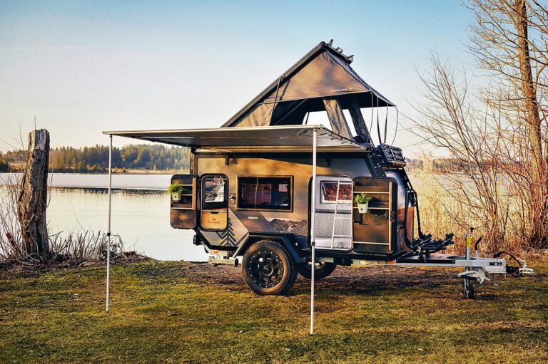 Ein robuster Offroad-Camper-Anhänger mit ausziehbarer Markise und Dachzelt steht direkt am Seeufer – umgeben von Wiese, kahlen Bäumen und ruhigem Wasser unter klarem Himmel. Dieser Custom Camper ist ideal für Abenteurer, die mit Pickup Camper oder Geländewohnwagen unabhängig unterwegs sein wollen und Wert auf Flexibilität sowie maximalen Komfort legen. Travelboxx - Pickup Wohnkabinen
