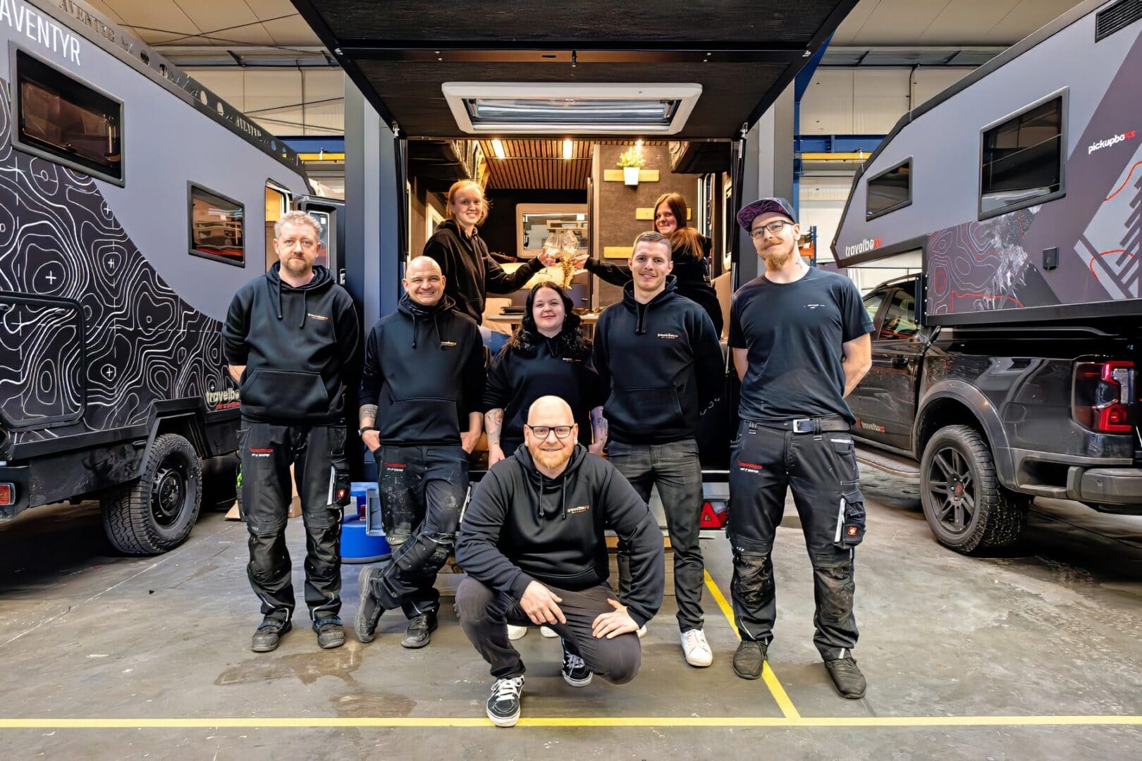 Neun Mitarbeitende in schwarzen Arbeitsoutfits lächeln vor einer Werkstatt mit zwei Offroad-Campern und Pickup-Campern. Travelboxx - Tiny House Wohnkabinen