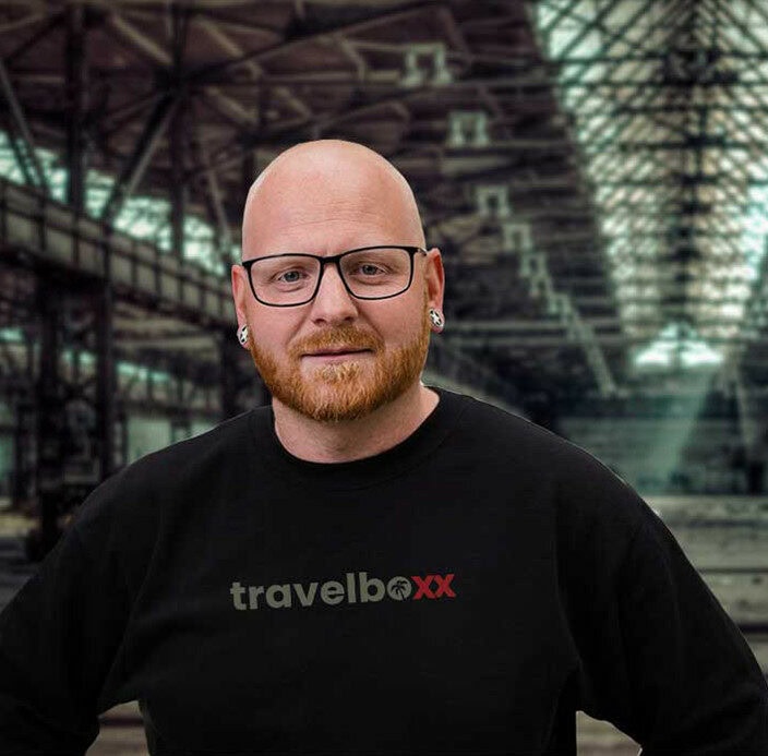 Benjamin Benny Baier mit einem schwarzen „travelboxx“ Sweatshirt steht vor einer großen Industriehalle mit Glasdach – perfekt für alle, die sich für Pickup Camper, Offroad Camper oder individuell angepasste Custom Camper interessieren. Travelboxx - Pickup Wohnkabinen